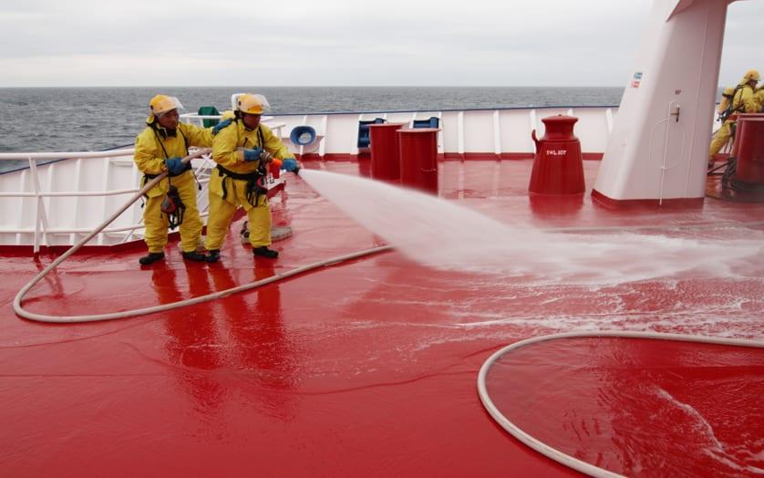 Firefighters in yellow suits using water hose on red deck of a marine vessel at sea