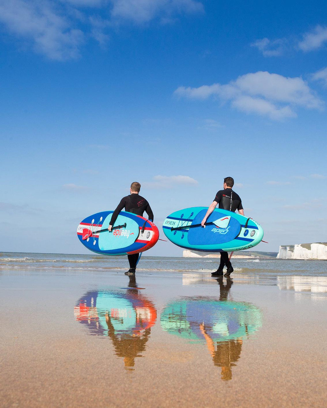 Two people in wetsuits carrying paddle boards on a beach near the ocean, with blue sky above.