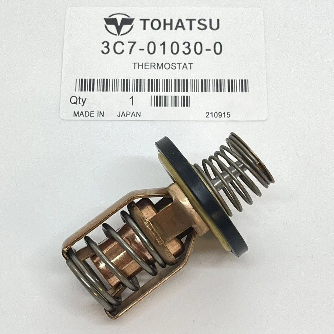 A Tohatsu thermostat part, model 3C7-01030-0, shown on a white background with its original product label card. The thermostat has a brass-colored body with internal springs and a black rubber gasket.